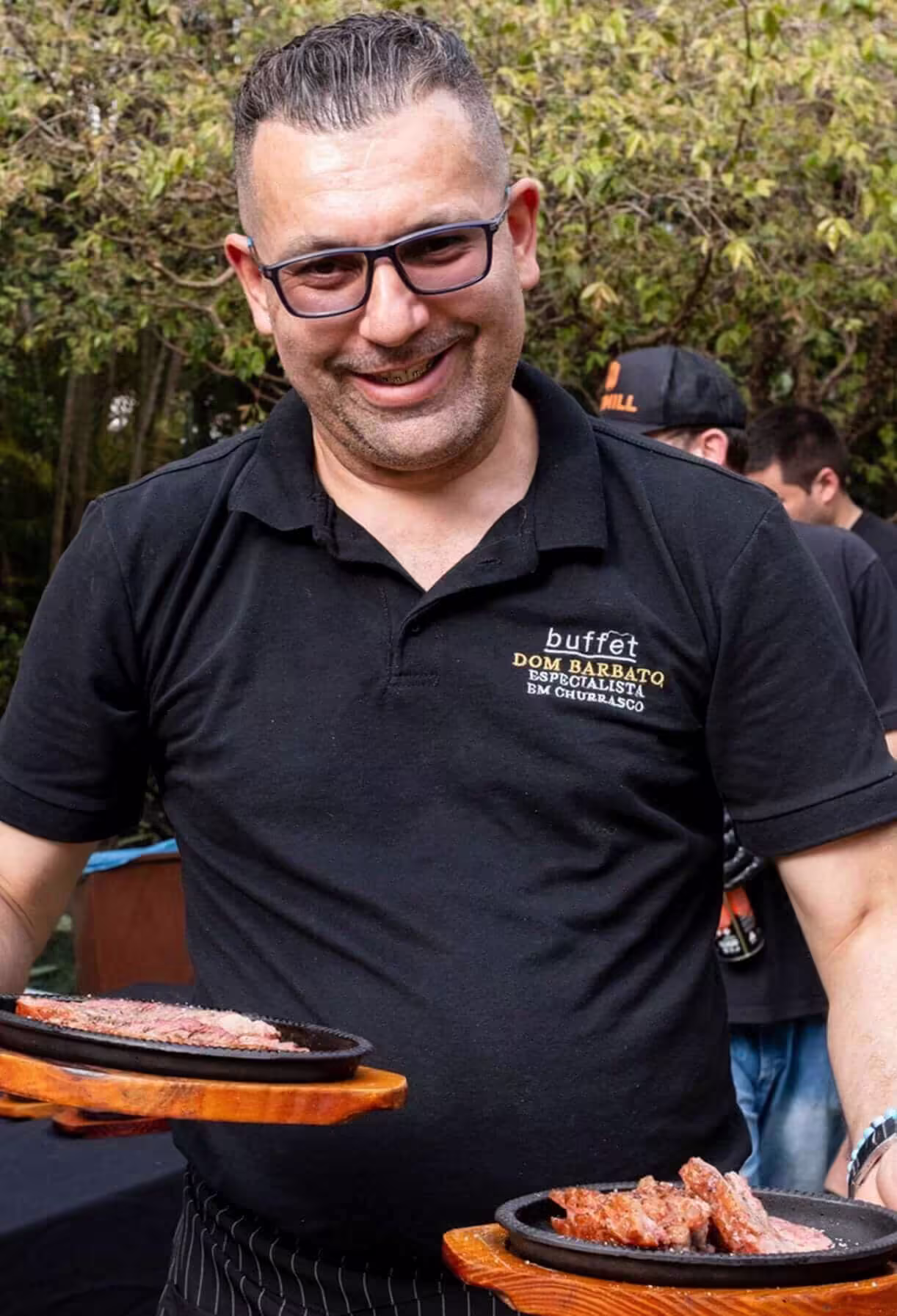 Chef Dom Barbato sorridente apresentando dois pratos de carne grelhada em tábuas de madeira