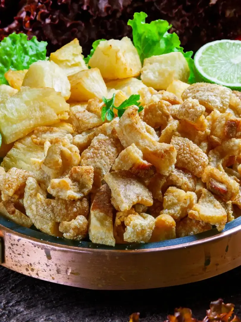 Prato com torresmo frito dourado, mandioca cozida em cubos, folhas de alface e fatia de limão