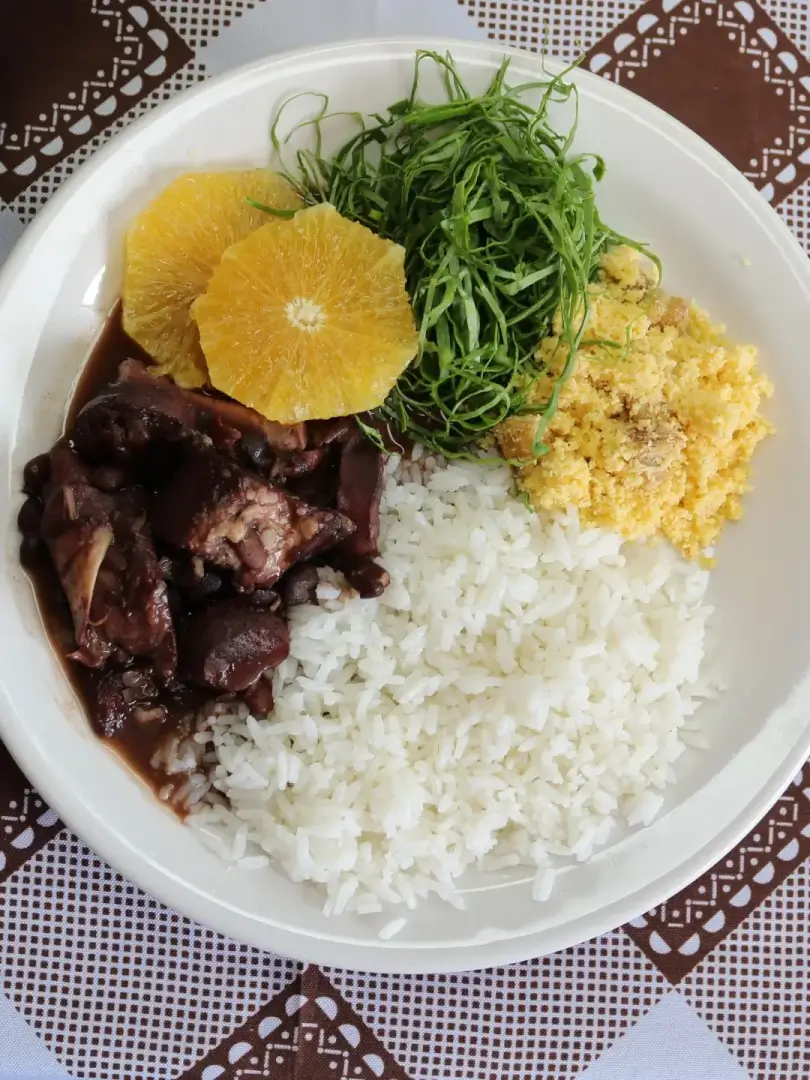 Prato de feijoada com arroz branco, farofa amarela, couve fatiada fina e fatia de laranja sobre prato branco