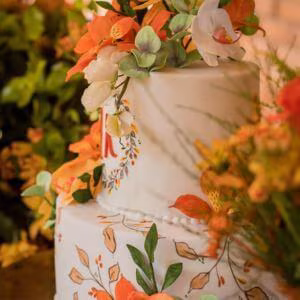 Bolo de casamento branco de três andares decorado com flores laranja e brancas e detalhes pintados à mão