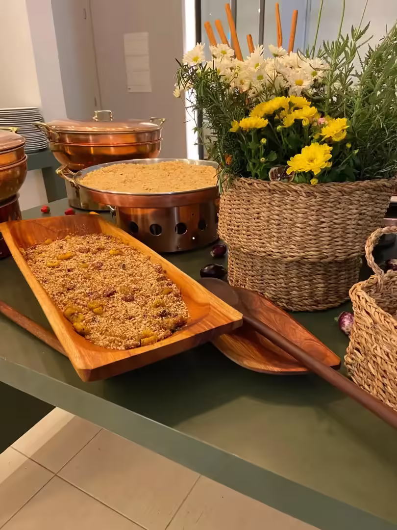 Bandeja de madeira alongada com farofa e frutas secas ao lado de arranjo de flores amarelas e brancas em cesto de palha