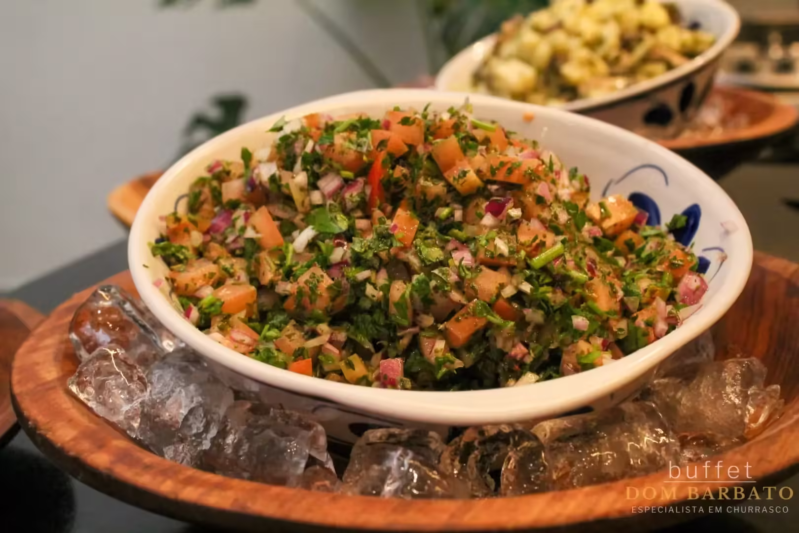 Tigela branca com salada picada verde e vermelha sobre base de madeira com gelo, pratos ao fundo