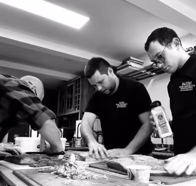 Três chefs em cozinha profissional preparando carne sobre bancada de madeira com facas e ingredientes