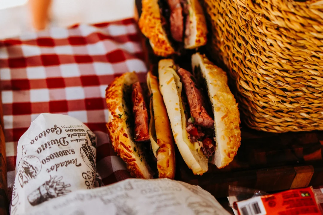 Três sanduíches artesanais com carne fatiada e queijo derretido sobre guardanapo xadrez vermelho e branco, ao lado de cesta de vime