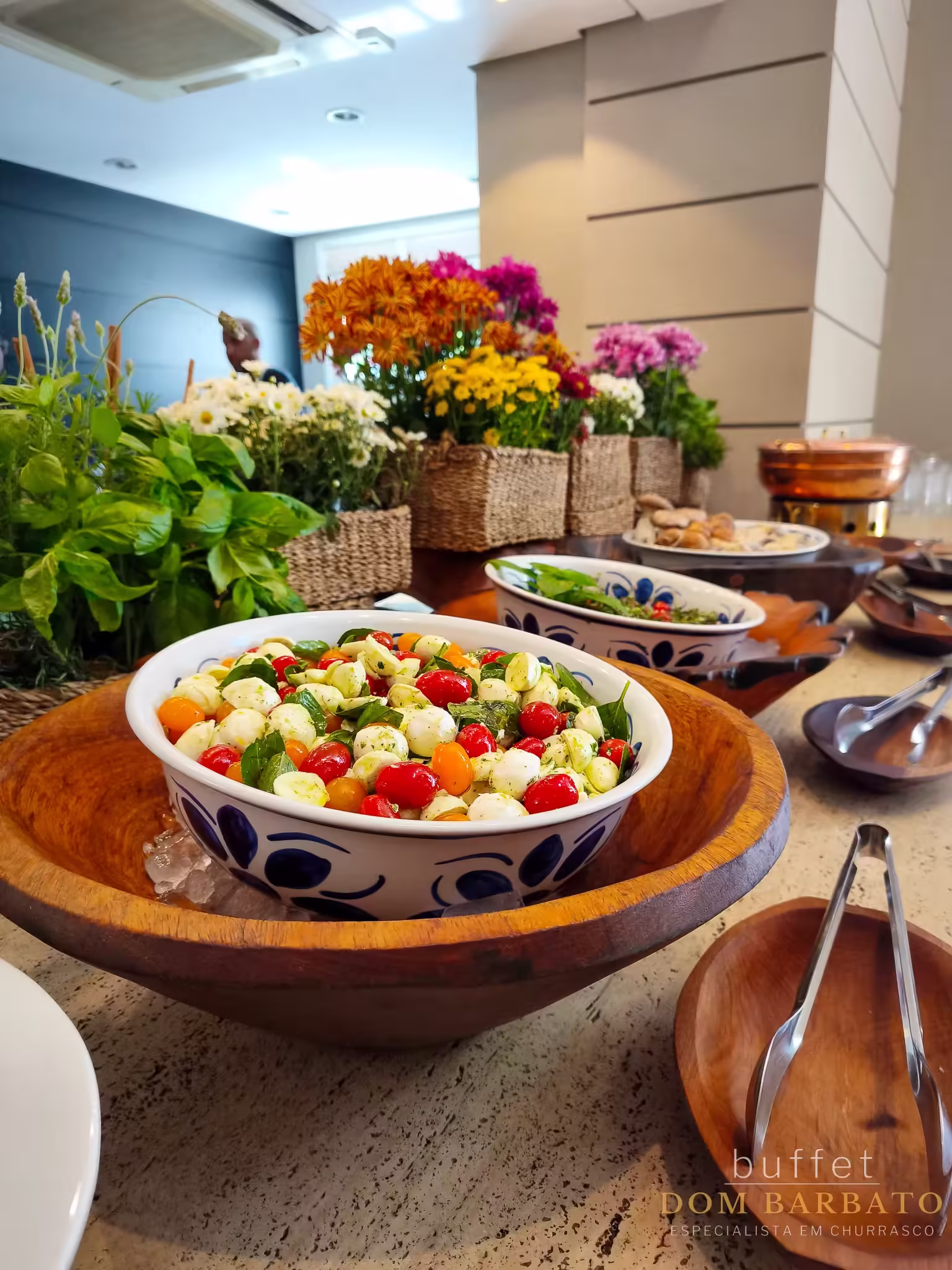 Bowl branco com detalhes azuis contendo salada de tomate-cereja, mussarela de búfala e folhas verdes, sobre tigela de madeira, em mesa de buffet com flores coloridas ao fundo