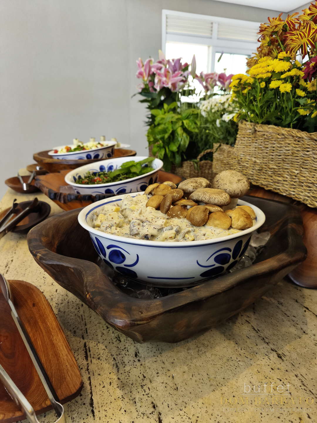 Bowl cerâmico azul e branco com salada de maionese e cogumelos frescos, sobre suporte de madeira rústico e flores ao fundo