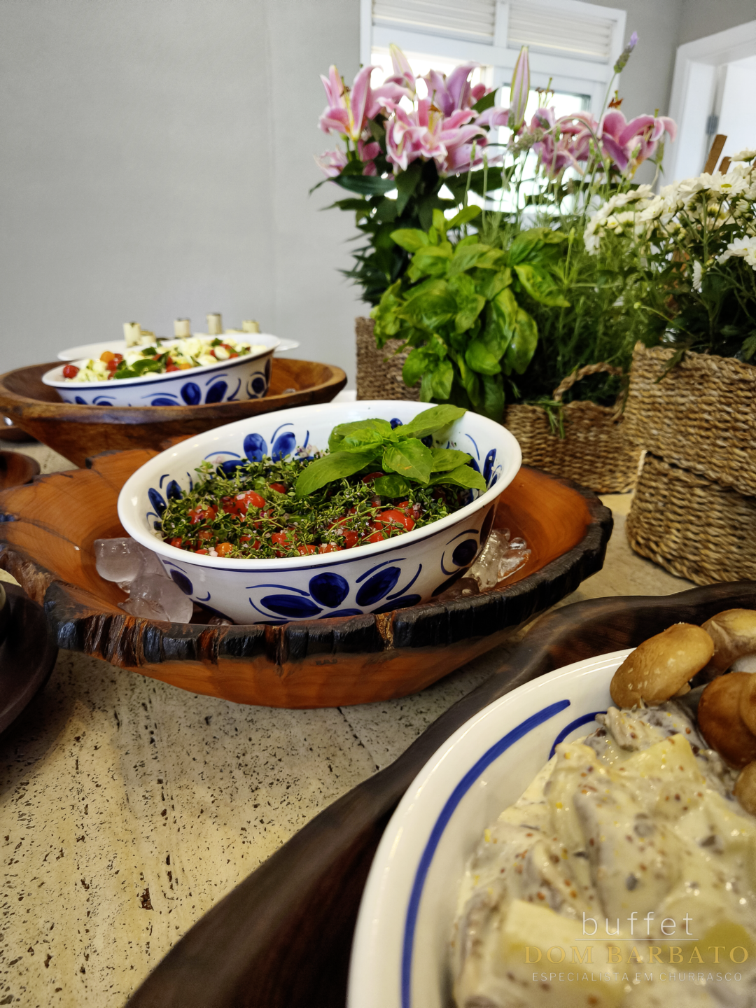 Mesa com potes de cerâmica azul e branca contendo saladas, tomate cereja e ervas, cercados por flores e cestos de vime