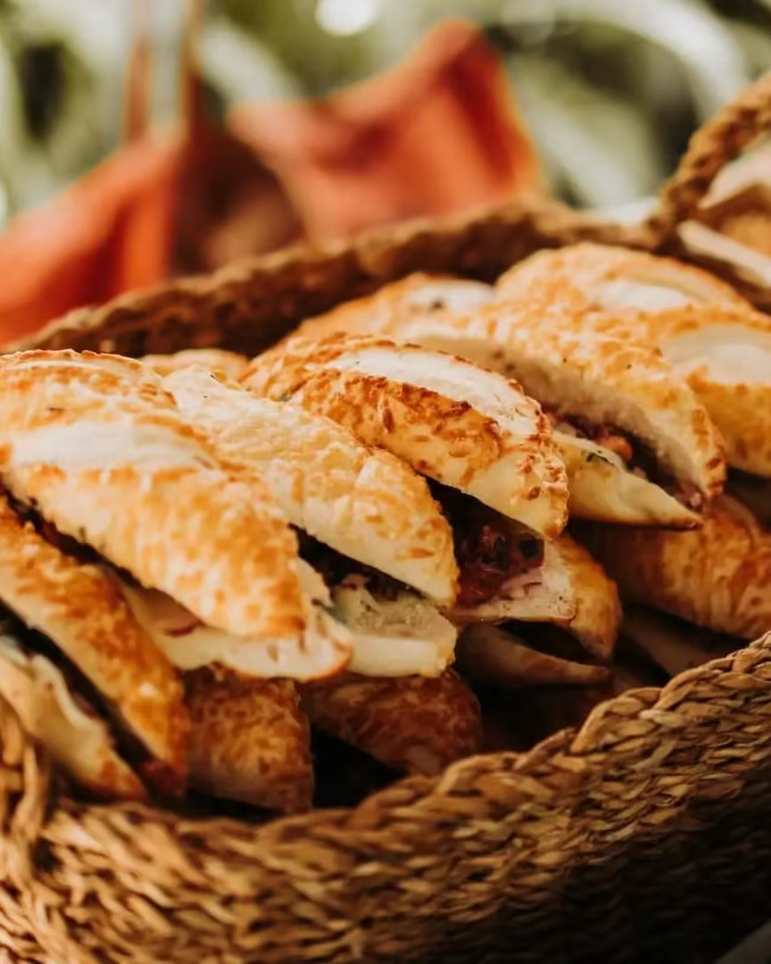 Cesta de vime com vários pães tipo baguete recheados e tostados, sobre toalha xadrez vermelha e branca