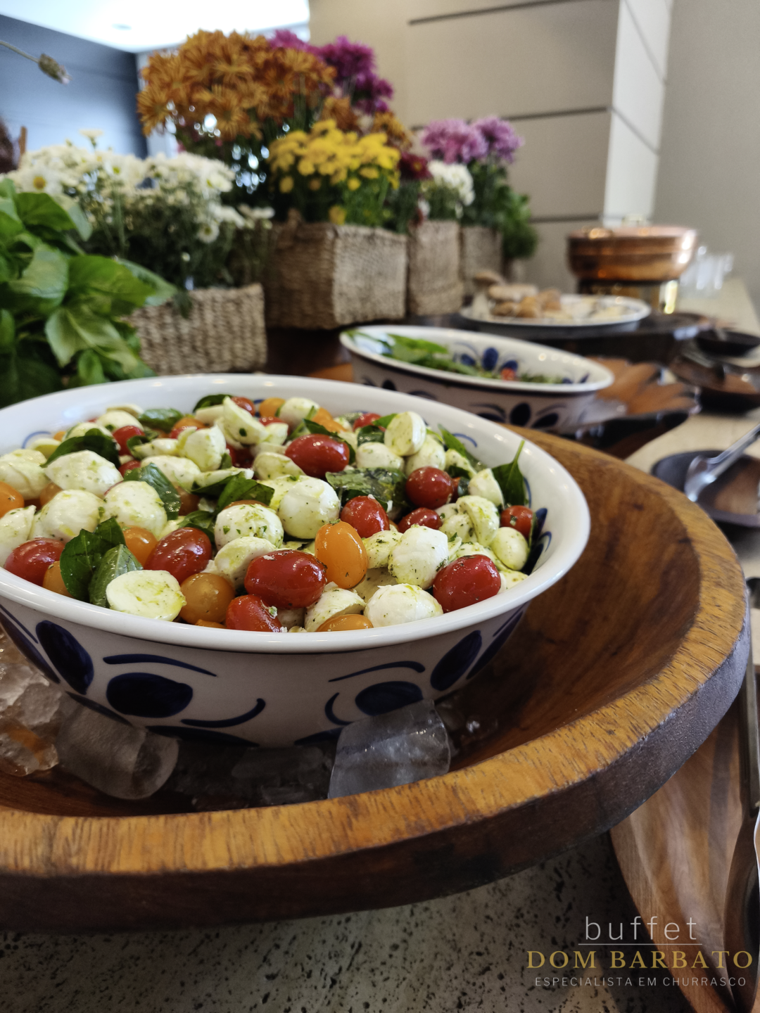 Tigela de cerâmica azul e branca com salada caprese de tomates cereja e mussarela, flores ao fundo
