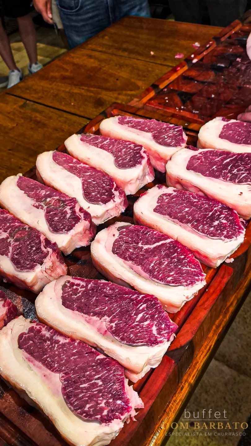 Cortes de carne crua com generosa camada de gordura dispostos em fileiras sobre tábuas de madeira em mesa