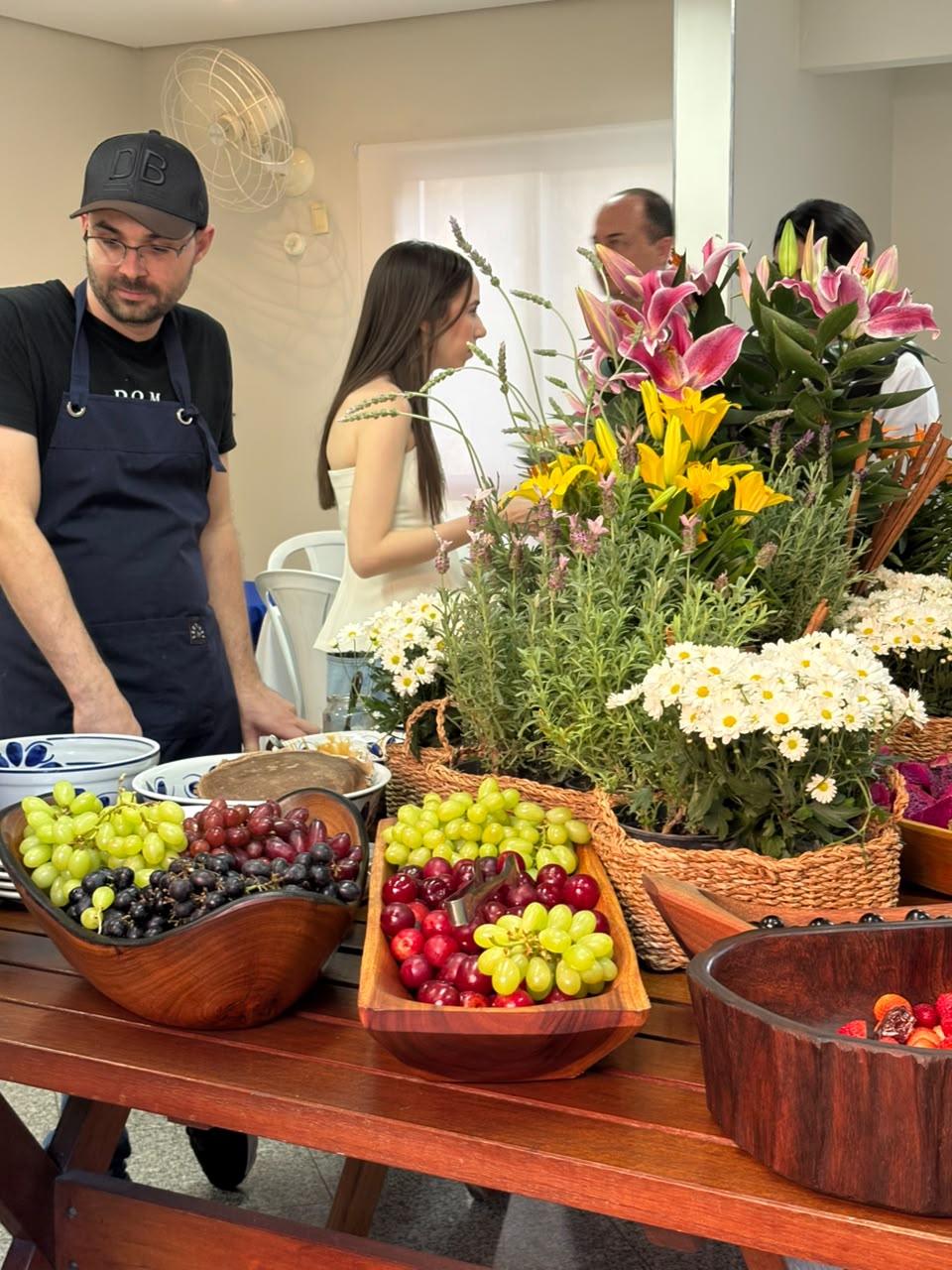 Mesa com tigelas de madeira contendo uvas verdes e roxas, cerejas, ao fundo arranjo de flores e pessoas
