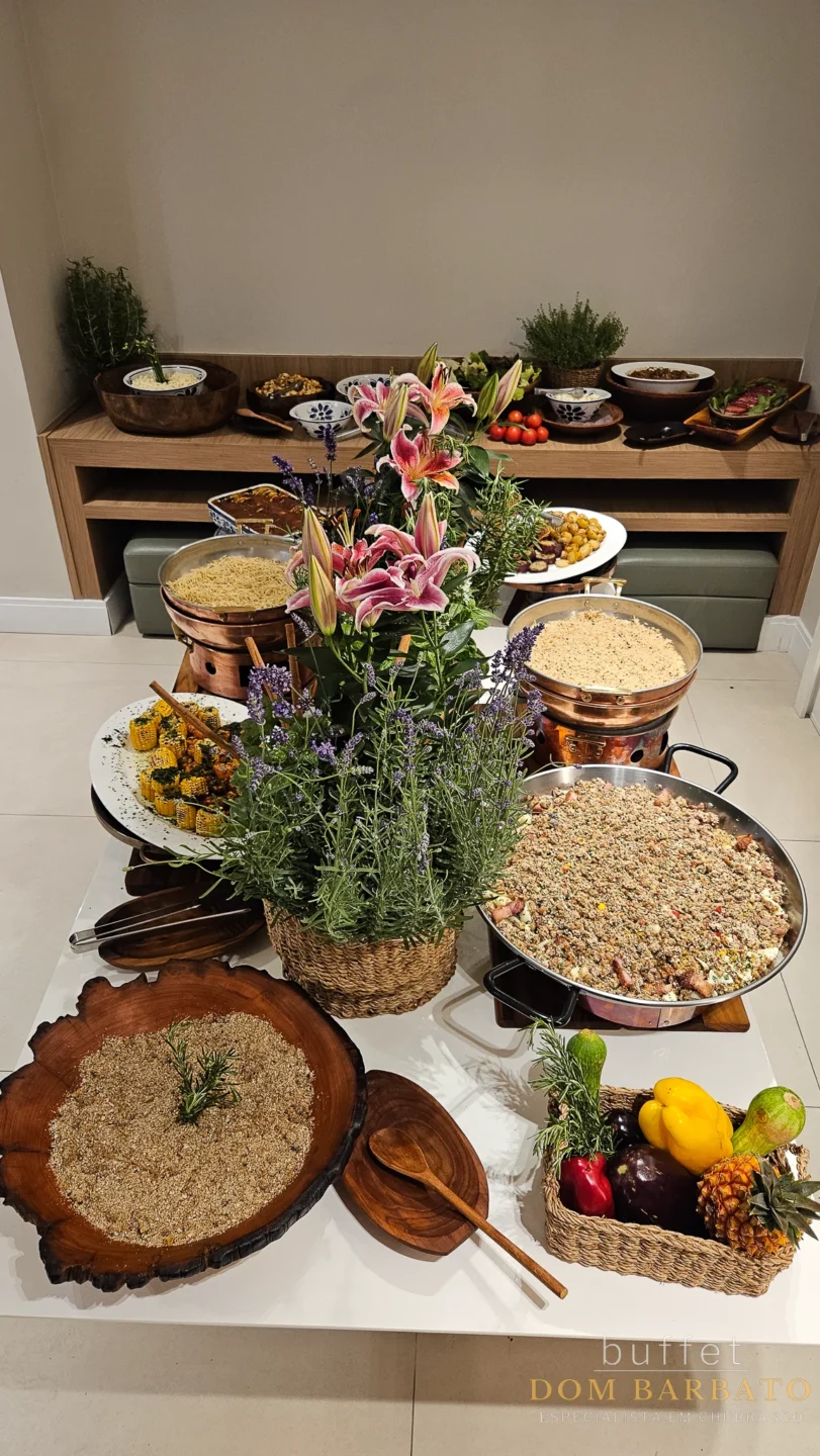 Buffet spread with multiple dishes, flowers, rice, grains, fresh vegetables, and herbs on white surface and wooden shelf backdrop