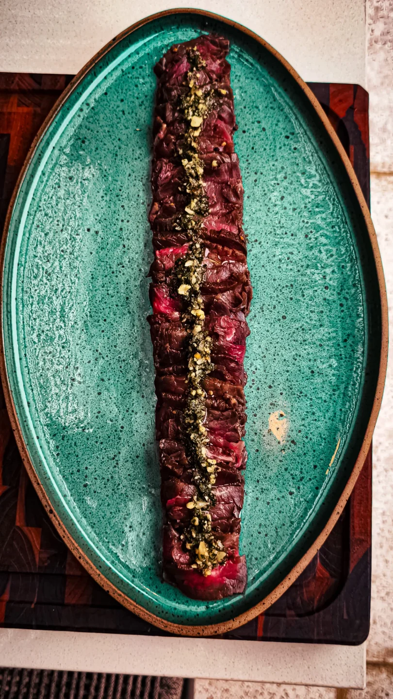 Fatias finas de carpaccio de carne em prato oval verde-água, decorado com chimichurri e castanhas picadas
