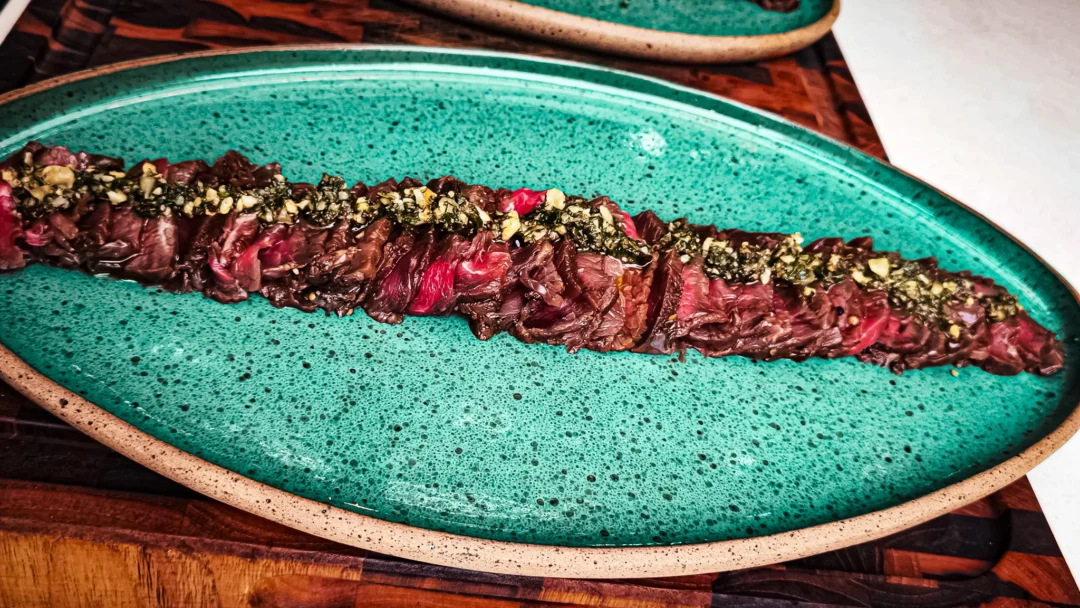 Fatias finas de carpaccio de carne dispostas em linha sobre prato oval verde-turquesa, cobertas com chimichurri e castanhas picadas