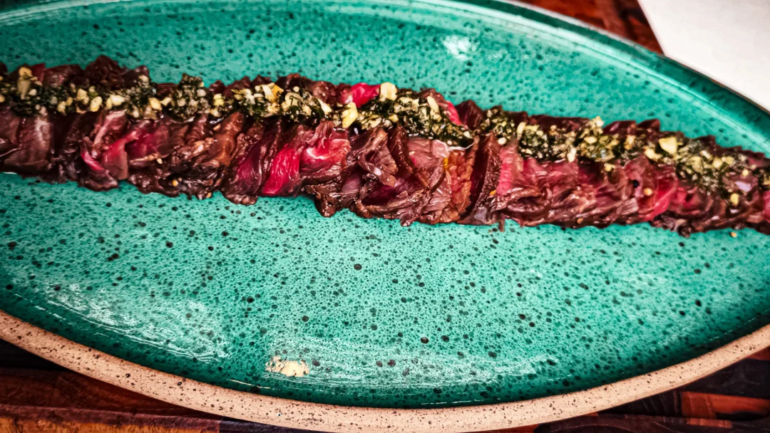 Fatias finas de carpaccio de carne dispostas em linha sobre prato oval verde-turquesa, cobertas com chimichurri e castanhas picadas