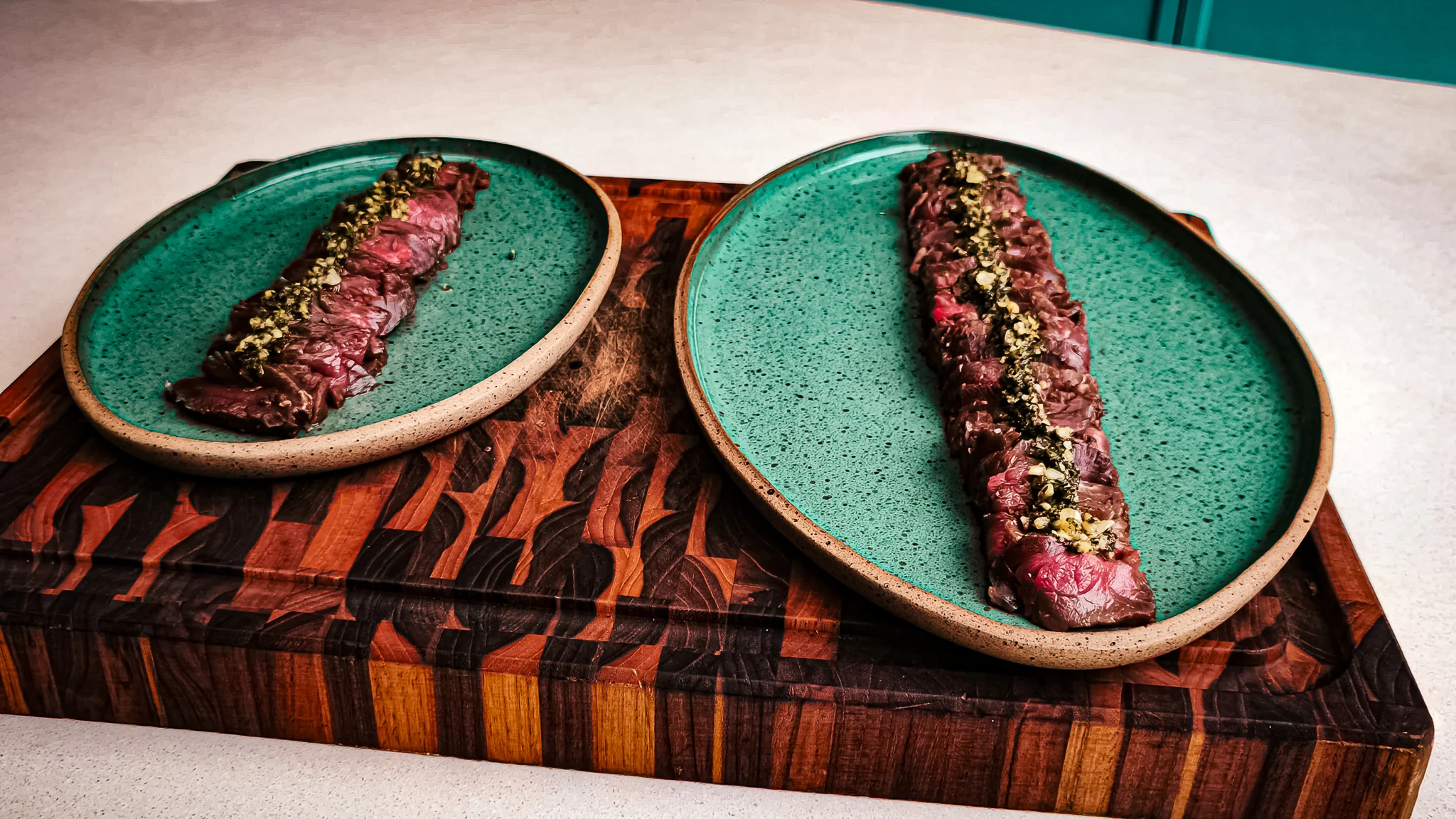 Duas fatias longas de carpaccio de carne cru com chimichurri e castanhas em pratos verdes sobre tábua de madeira