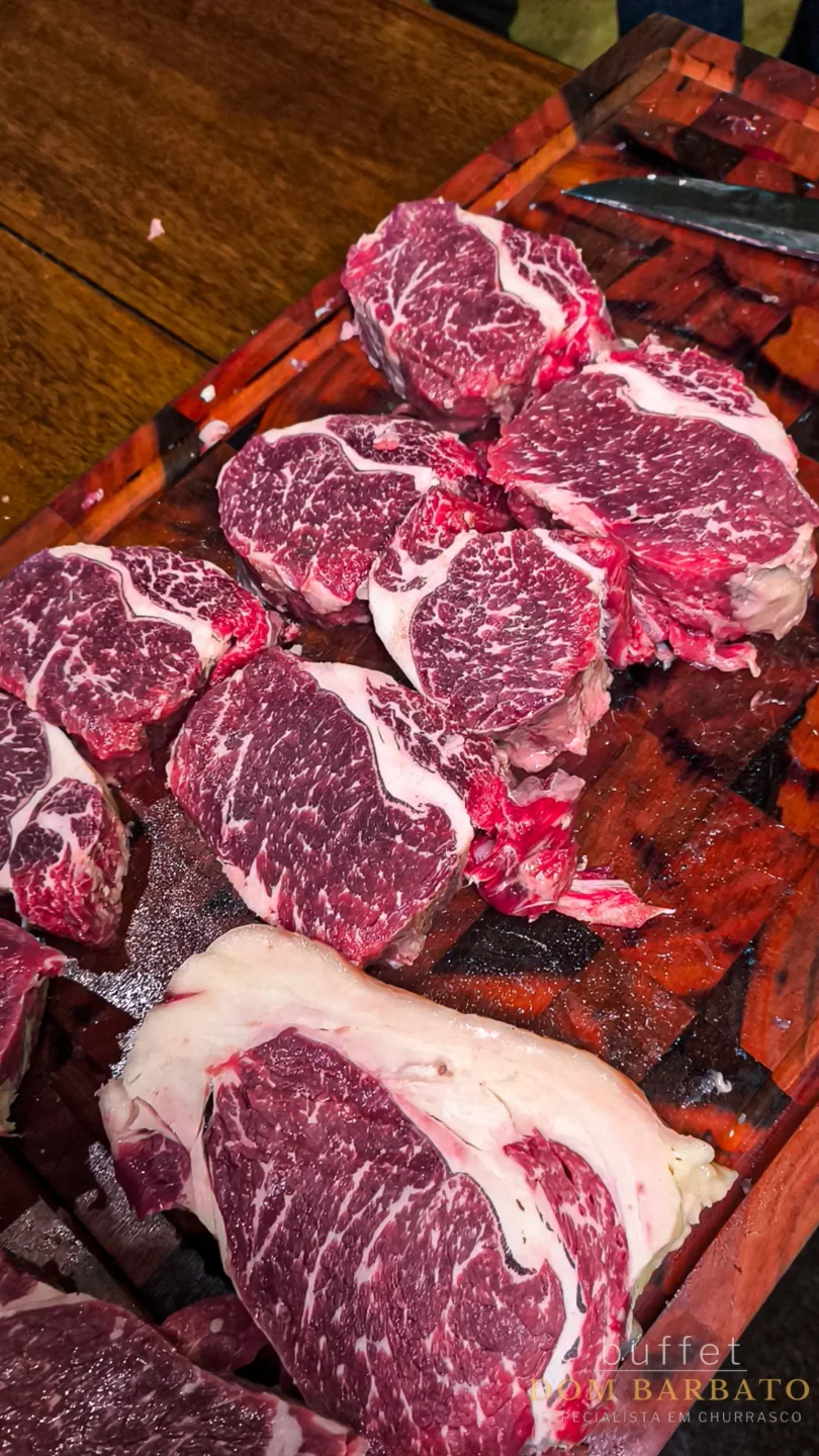 Cortes de carne bovina crua com marmoreio sobre tábua de madeira escura, faca ao fundo