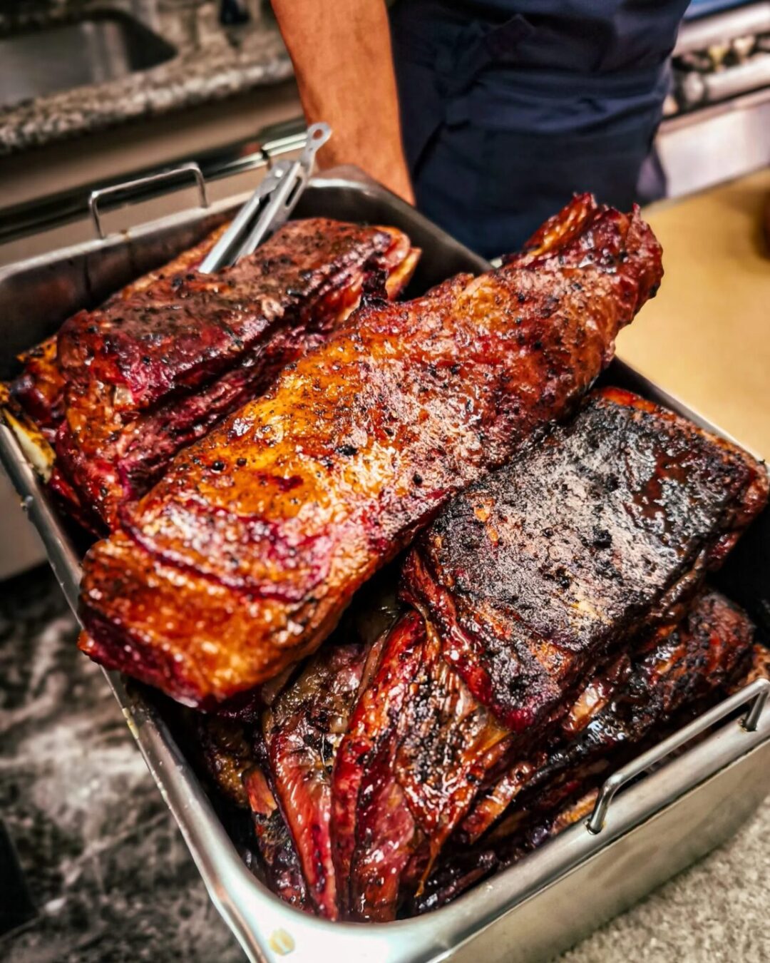 Bandeja de inox com grandes peças de carne assada escura, segurada por homem de avental