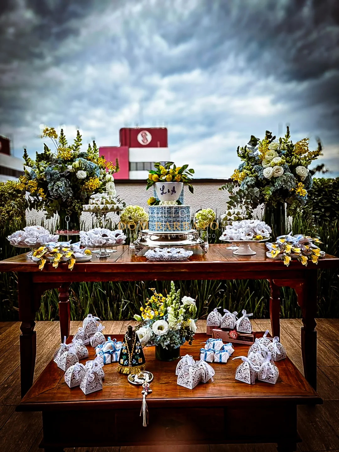 Mesa de madeira com bolo azul e branco decorado com limões, arranjos florais brancos e amarelos, doces e lembrancinhas rendadas
