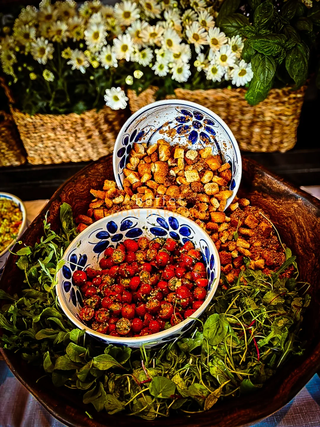 Tigela de madeira com croutons, tomates temperados em pratos de cerâmica azul e branca, ervas frescas e flores brancas ao fundo