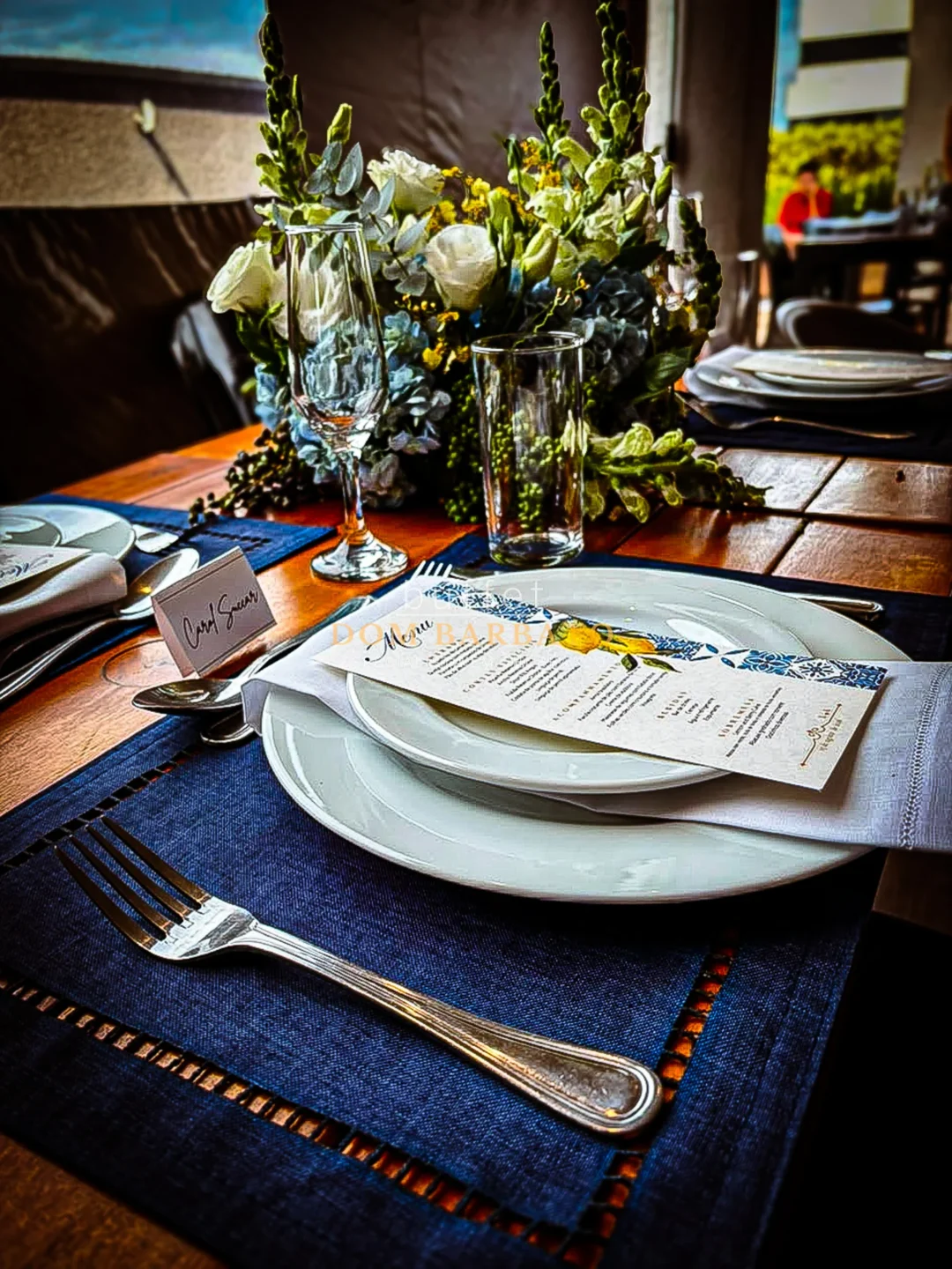 Mesa posta com pratos brancos, taça, guardanapo, cartão de nome manuscrito e arranjo de flores brancas ao fundo