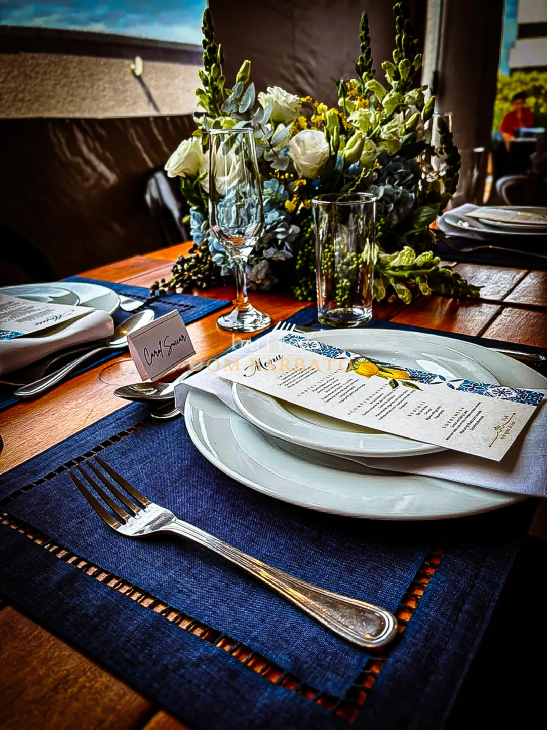 Mesa posta com pratos brancos, guardanapo, garfo sobre lugar americano azul-marinho e arranjo de flores brancas