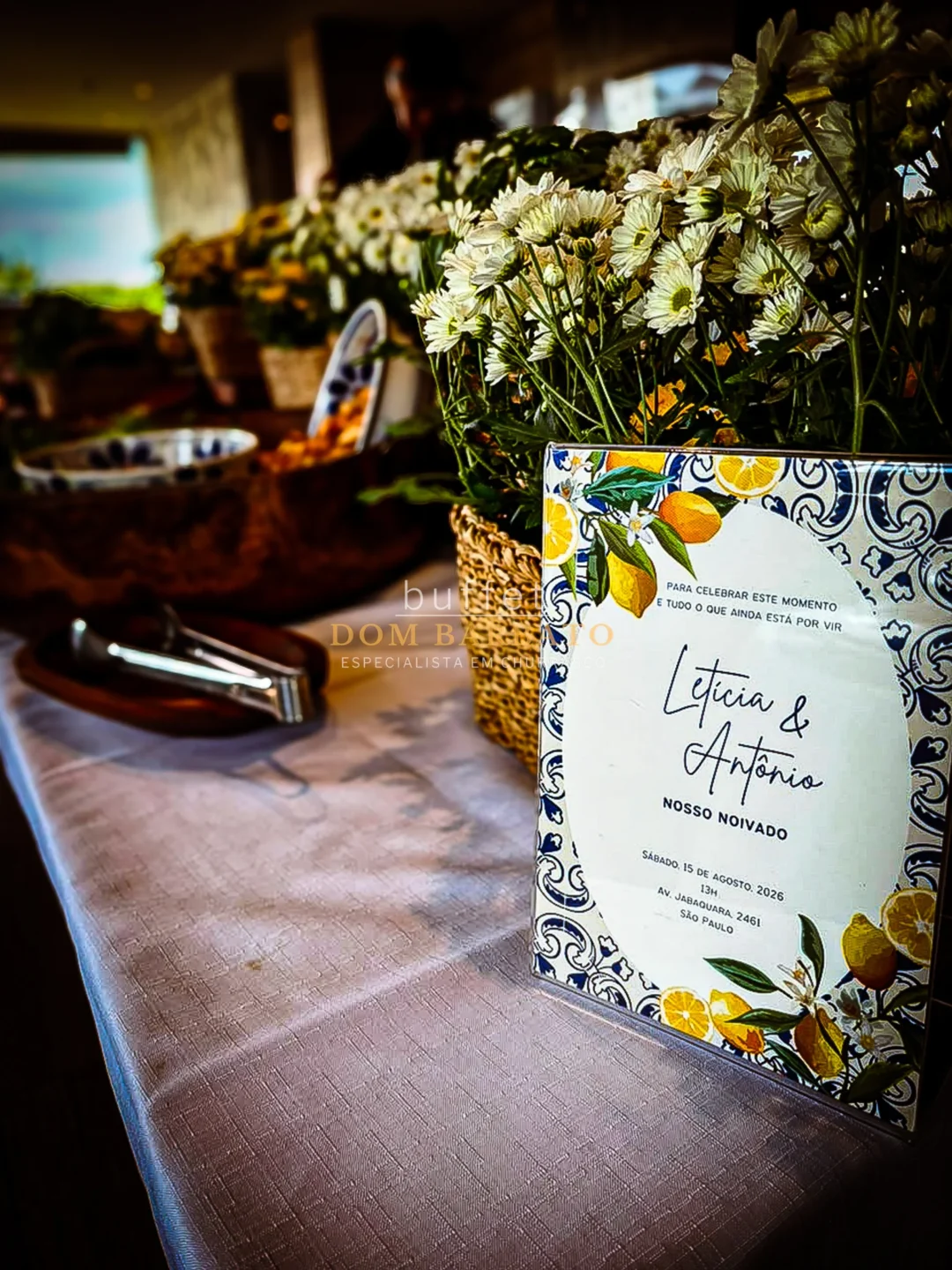 Placa decorativa com padrão azulejo azul e branco e ilustrações de limão siciliano, texto 'Letícia & Antônio Nosso Noivado', cercada por flores brancas em vasos de fibra natural sobre mesa com toalha rosa claro