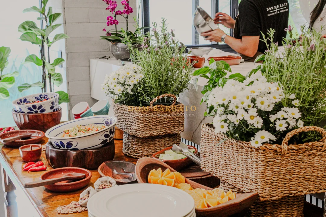 Mesa de buffet com tigelas de comida, flores brancas em cestos de fibra, pessoa ao fundo servindo