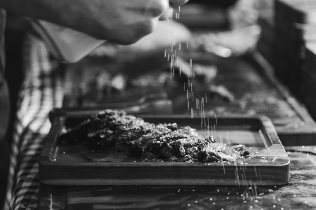 Mãos finalizando corte de carne grelhada em tábua de madeira, salpicando tempero granulado