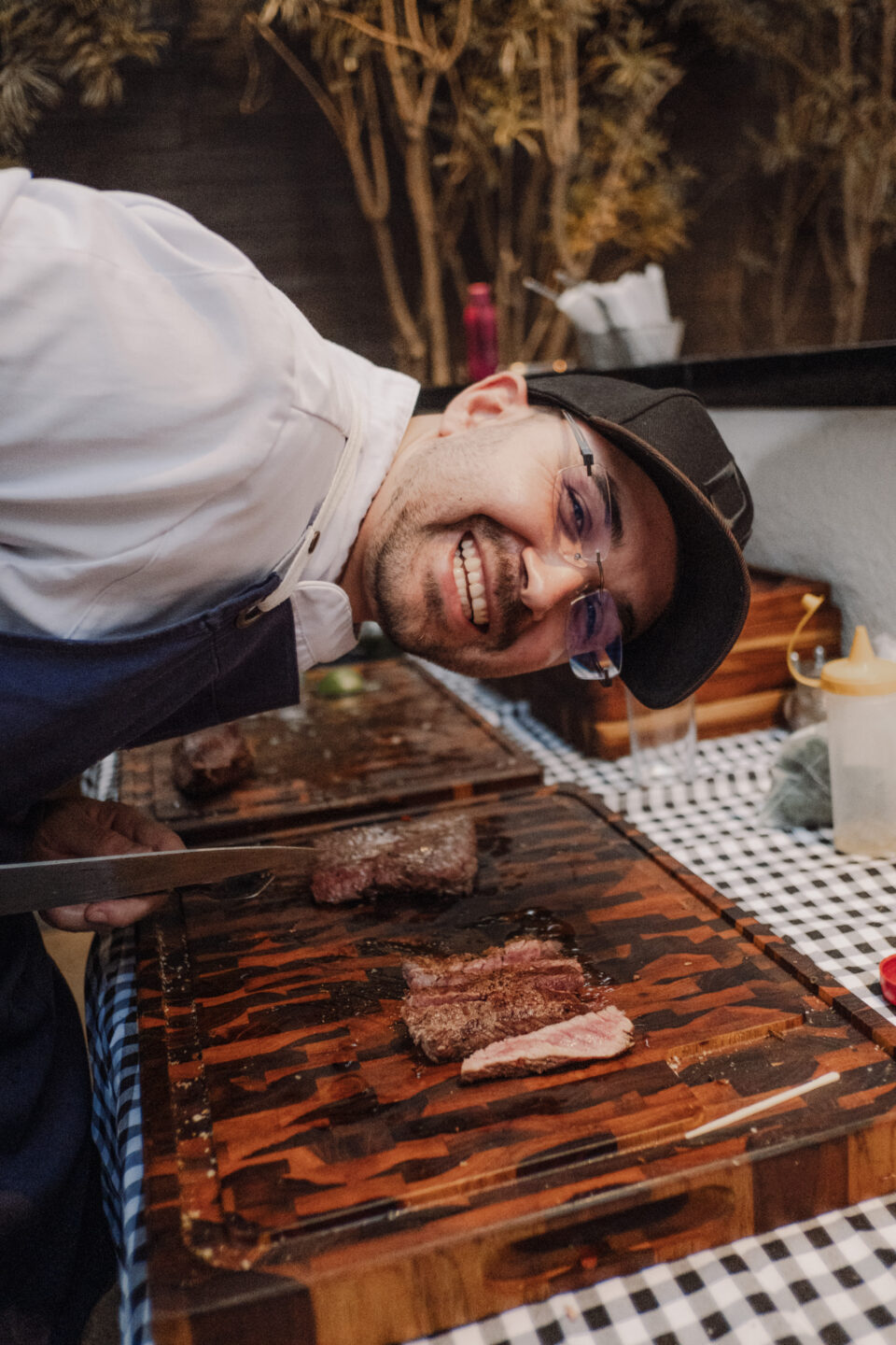 Chef sorridente com boné e óculos fatiando carne grelhada em tábua de madeira