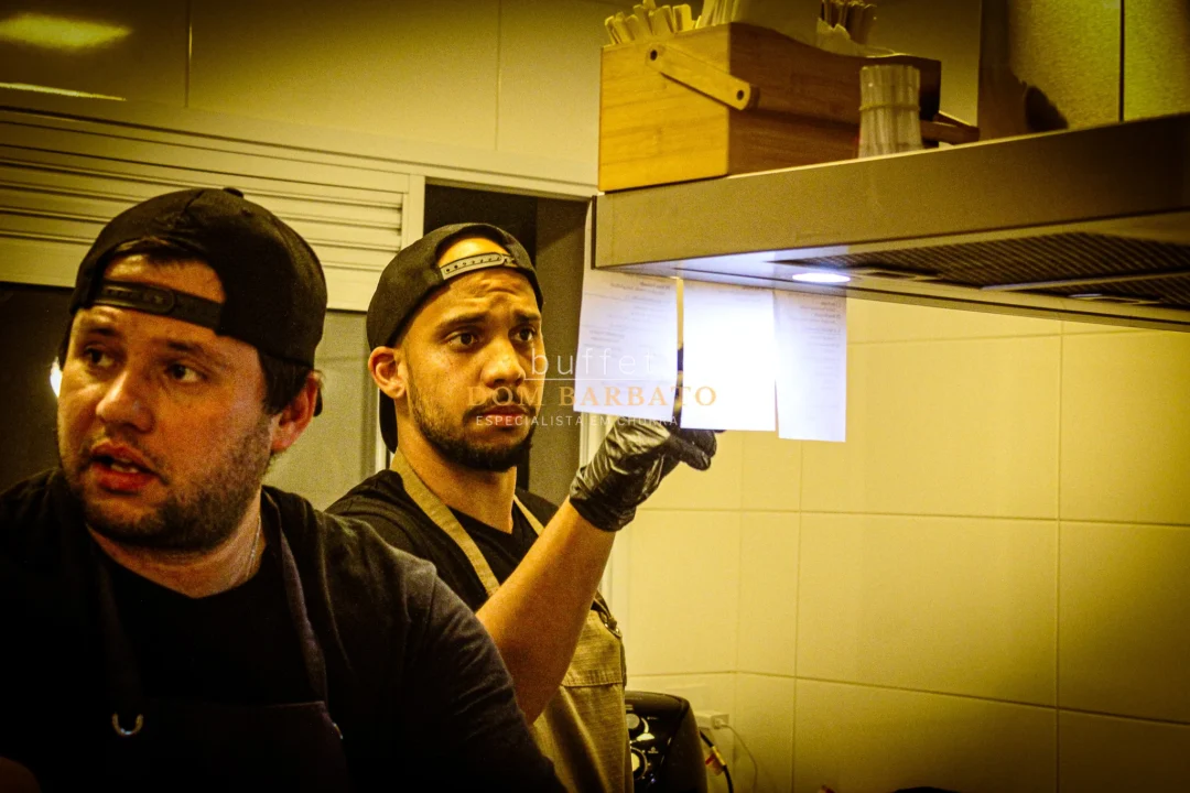 Dois cozinheiros de boné preto na cozinha; um segura comanda de papel com luva.