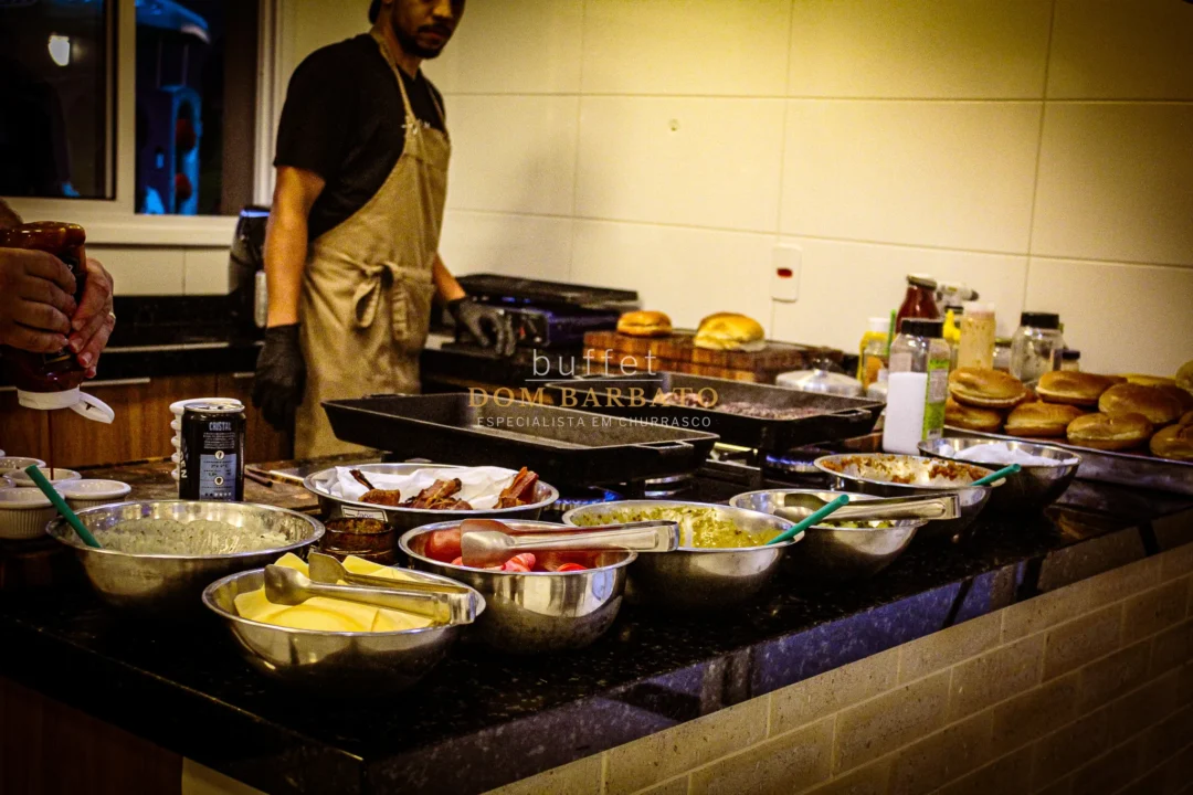 Balcão de buffet com tigelas de acompanhamentos, queijos, tomates e pães ao fundo com cozinheiro de avental.