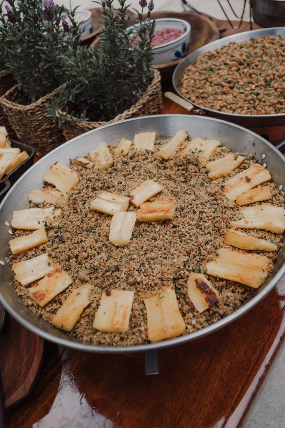 Bandeja redonda com farofa e tiras de mandioca grelhada dispostas em círculo, vasos de alecrim ao fundo