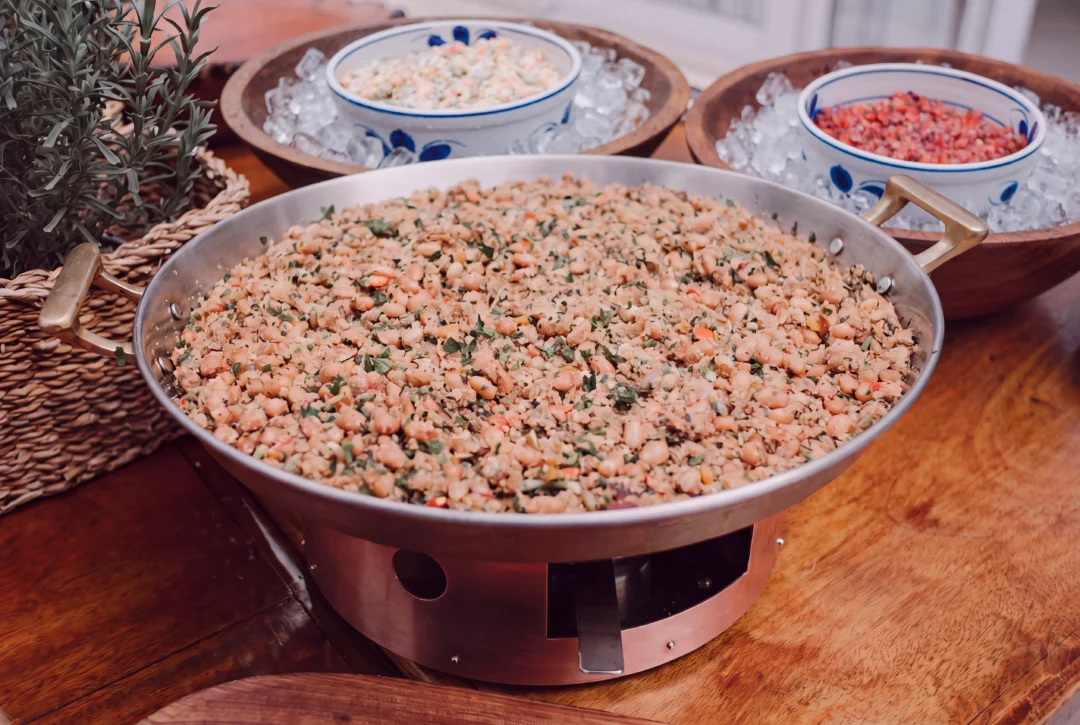 Panela grande de cobre com feijão tropeiro guarnecida com ervas, ao lado de tigelas de porcelana com maionese de legumes e vinagrete sobre gelo, em mesa de madeira com planta ao fundo