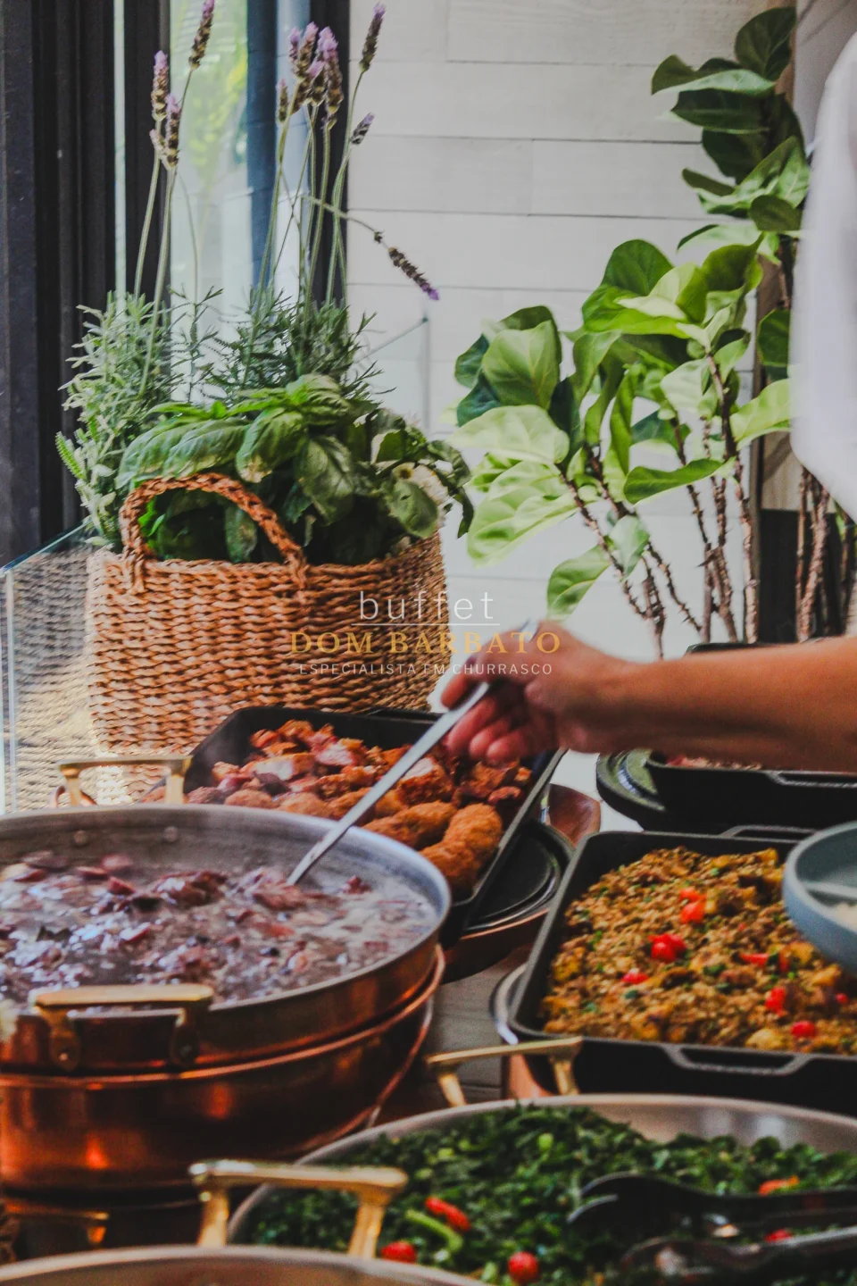 Panela de cobre com feijoada, bandejas de carnes, couve e arroz em buffet self-service com cesta de temperos frescos