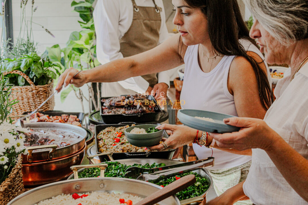 Mulher se serve em buffet com feijoada, arroz, couve e carnes, outra mulher segura tigela cinza