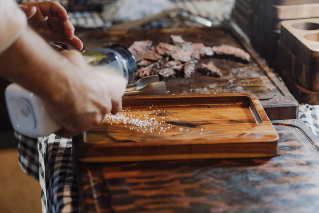 Mão finalizando corte de carne grelhada sobre tábua de madeira, salpicando sal grosso