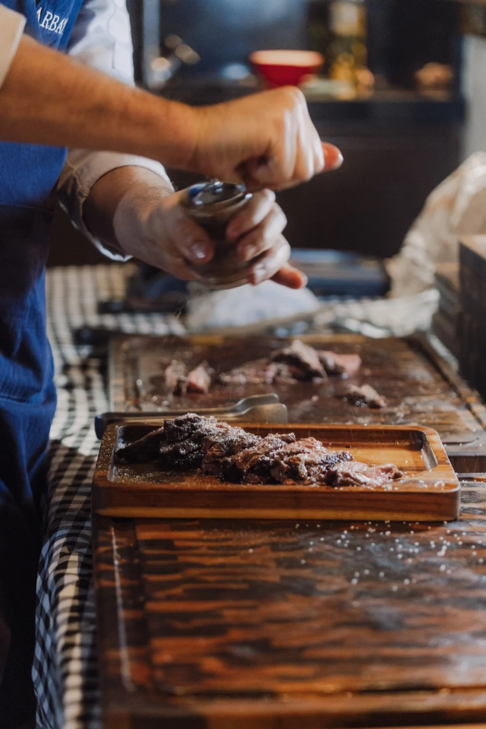 Churrasqueiro finaliza carne fatiada com sal grosso em tábua de madeira sobre bancada com toalha xadrez