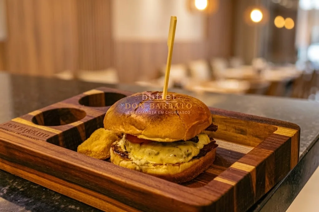 Hambúrguer com queijo derretido e espeto de madeira sobre tábua, com bolinho ao lado, em bar.