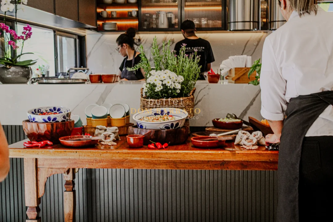 Mesa de madeira com tigelas de acompanhamentos, pimentas, flores e cozinheiros ao fundo em cozinha aberta