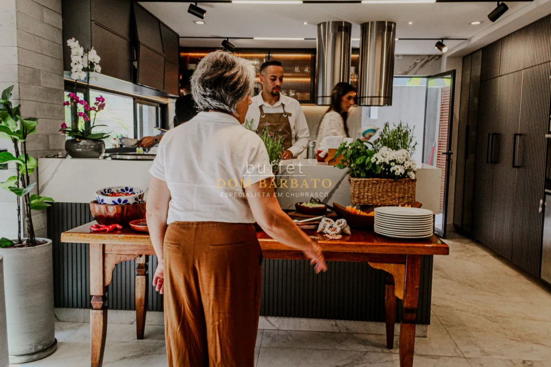 Mulher de costas observa mesa de madeira com pratos, flores e petiscos em cozinha aberta com chef ao fundo