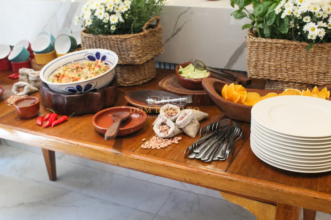 Mesa de madeira rústica com feijão em travessa, saquinhos de feijão crú, pimentas, laranjas cortadas, talheres e pratos brancos empilhados, com cestos de flores brancas ao fundo