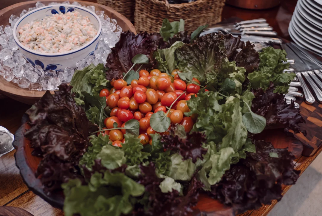 Bandeja com folhas de alface roxa e verde ao redor de tomates cereja, ao lado de salgada de bacalhau em bowl com gelo e talheres de prata