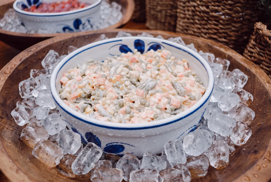 Tigela de porcelana com salada russa (maionese, ervilha, cenoura em cubos) sobre gelo, em bandeja de madeira