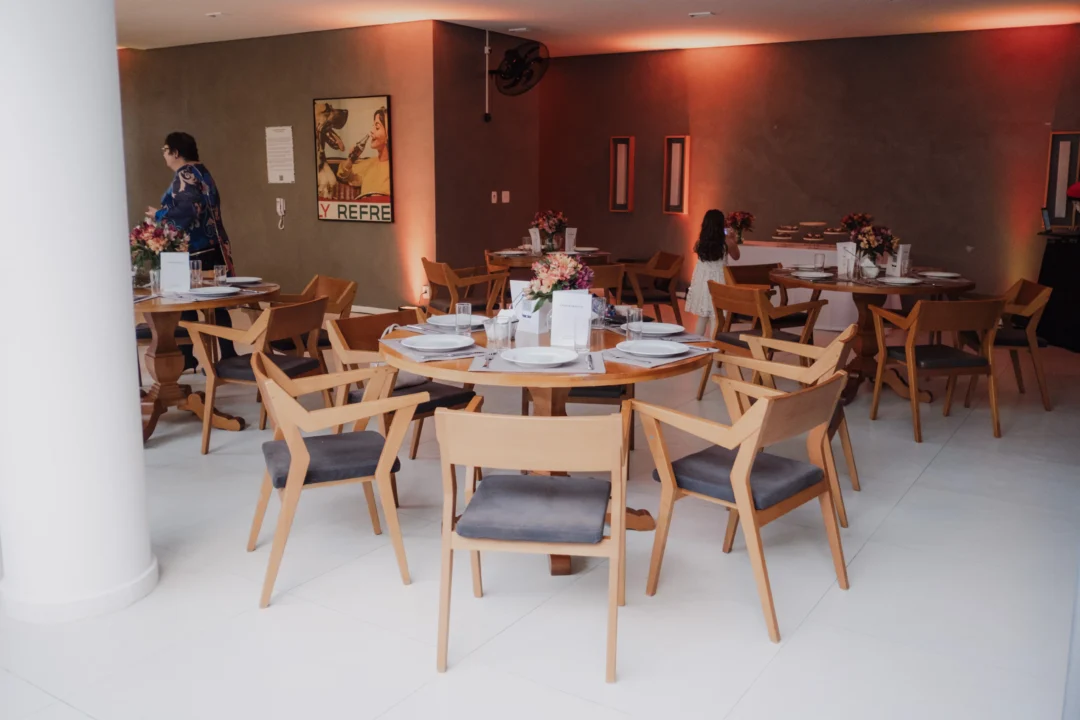 Salão decorado com mesas redondas de madeira, cadeiras estofadas, flores e louças brancas para evento