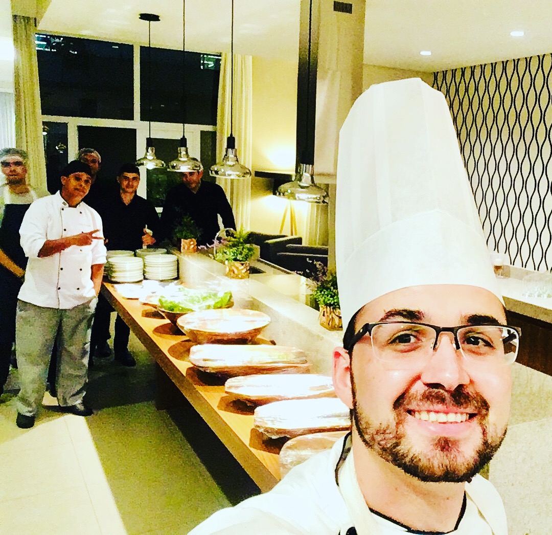 Chef de dólmã branca e touca alta sorri em selfie; equipe ao fundo junto ao balcão com pratos e travessas