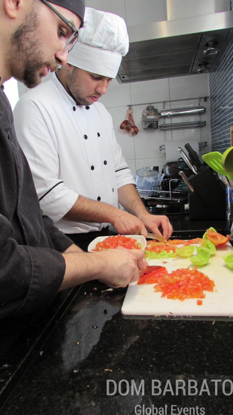 Dois homens cortam tomates em tábua branca na cozinha; um veste dólmã e chapéu de chef