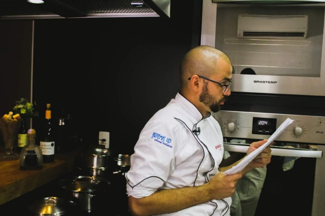 Chef de óculos e dólmã branca lê folhas de papel em cozinha com forno inox