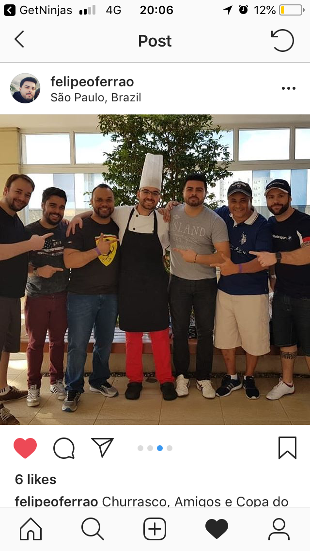 Seis homens apontam para chef de touca branca e avental preto diante de janelas e árvore
