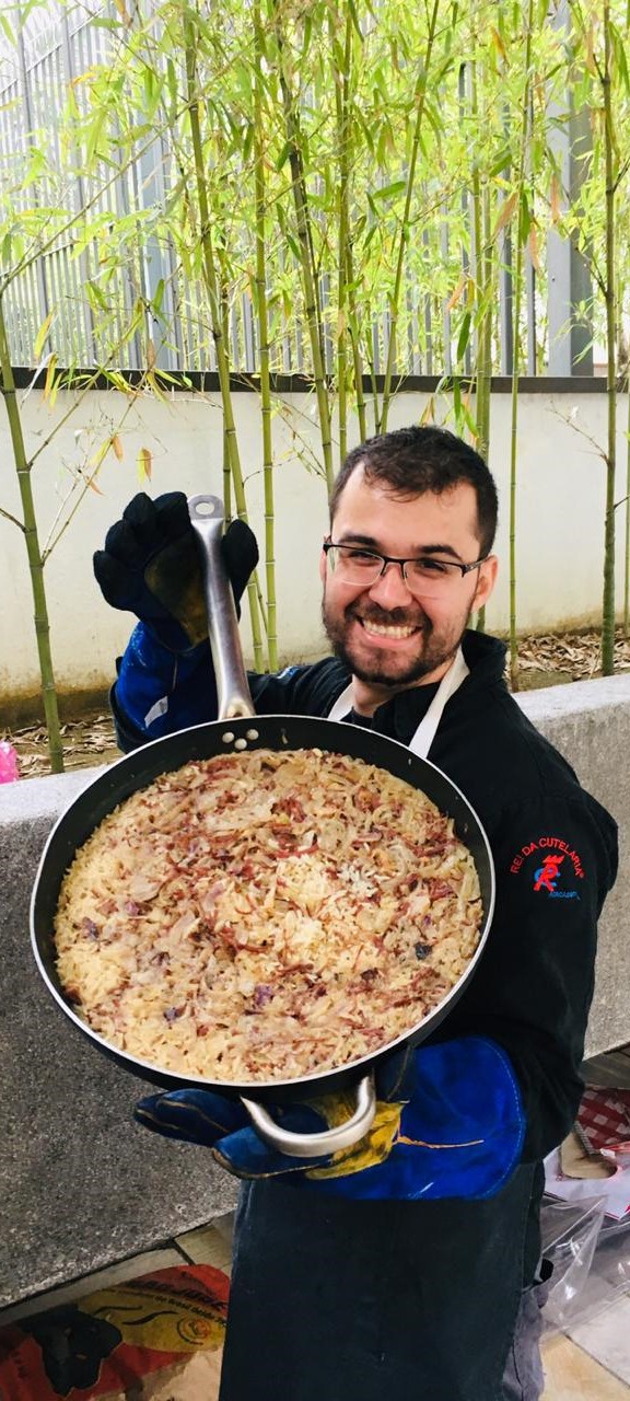 Homem de óculos e avental segura frigideira grande com arroz, cebola e carne, sorrindo ao ar livre